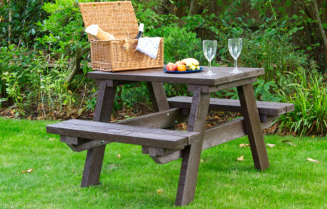 Small brown picnic table on grass with a picnic hamper, food and wine glasses set on it